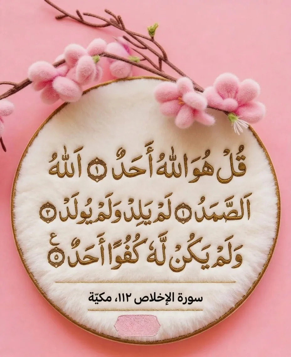 A close-up photograph of a round, gold-rimmed embroidery hoop holding a panel of fluffy, soft white plush fabric. Stretched over a flat, soft pink surface, the fabric is intricately embroidered with shimmering gold thread, rendering the full Arabic text of Surah Ikhlas (Surah 112) with ornate, framed-window style ayah markers. The detailed gold thread calligraphy is prominent and tactile. Above and to the left side of the hoop, a cluster of plush, soft pink cherry blossom-like flowers on branches are draped. Below the main Surah text, more gold thread embroidery provides the reference text: 'سورة الإخلاص ١١٢، مكية' (Surah Al-Ikhlas 112, Meccan). At the very bottom center of the hoop, a small, plain pink fabric patch is visible.