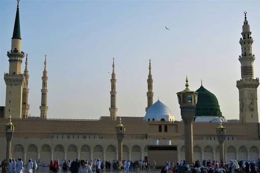 Exterior view of Al-Masjid an-Nabawi in Medina showing the iconic Green Dome, minarets, and worshippers gathering in the courtyard.